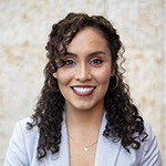 Veronica Arreola smiling, wearing a light gray blazer over a dark top, with shoulder-length dark curly hair, standing against a softly textured neutral background.