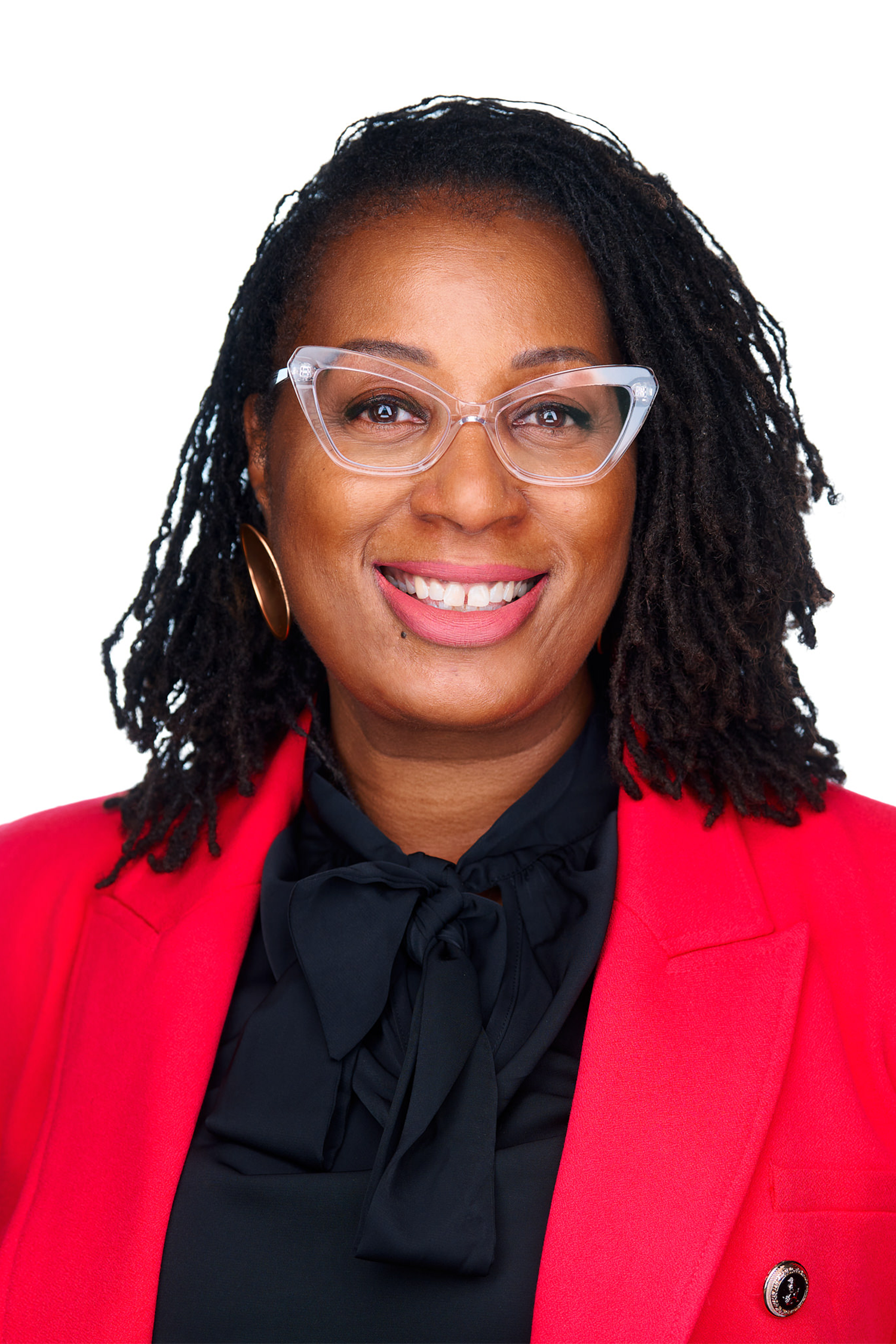 Latasha Smith, Ph.D., LCSW, CGP, smiling and wearing clear-framed glasses, a red blazer over a black blouse with a bow tie detail, and gold hoop earrings, against a light gray background.