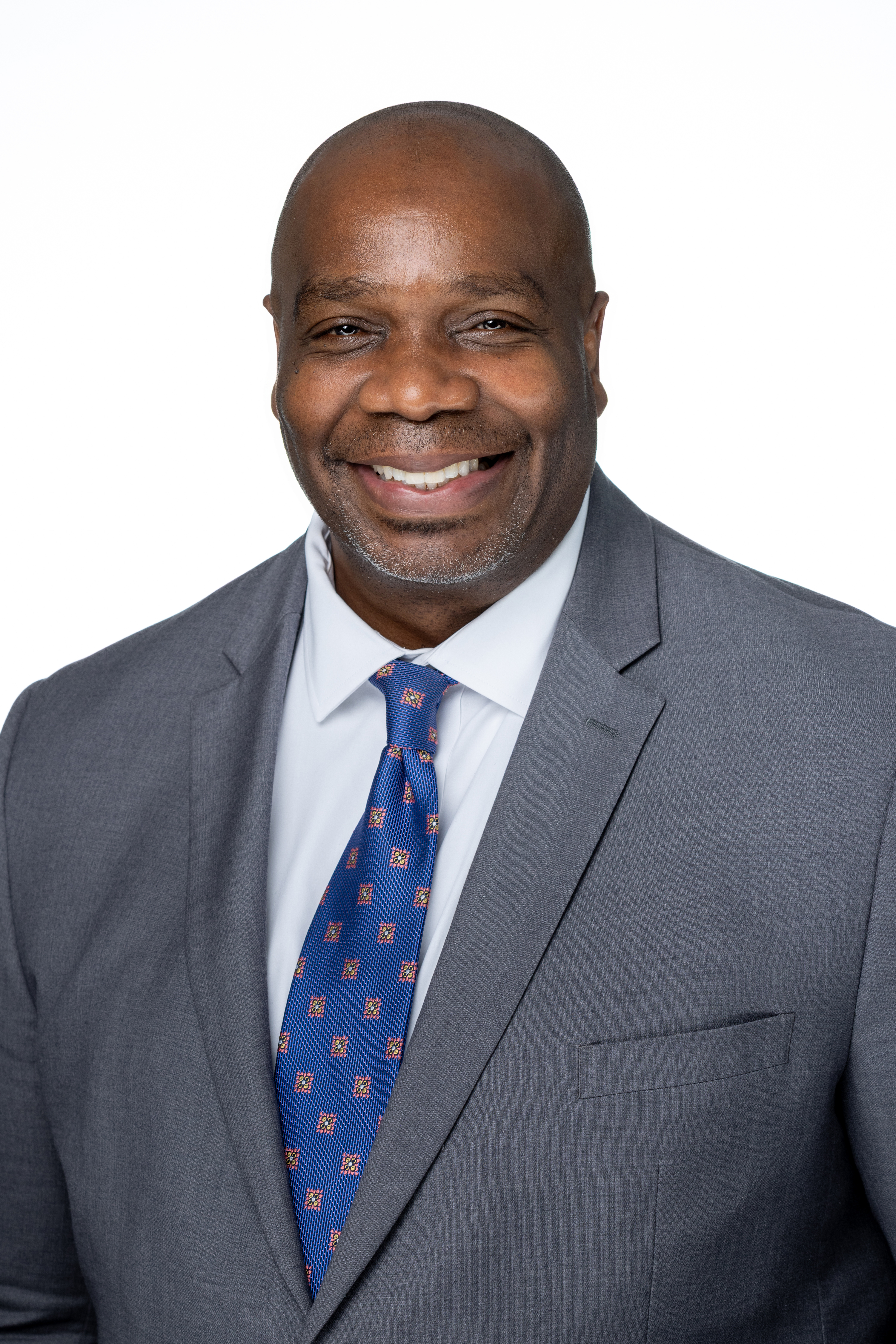 Amzie Moore II, a man with a shaved head and neatly trimmed facial hair, wearing a gray suit jacket, white dress shirt, and a blue patterned tie, smiling against a light gray background.