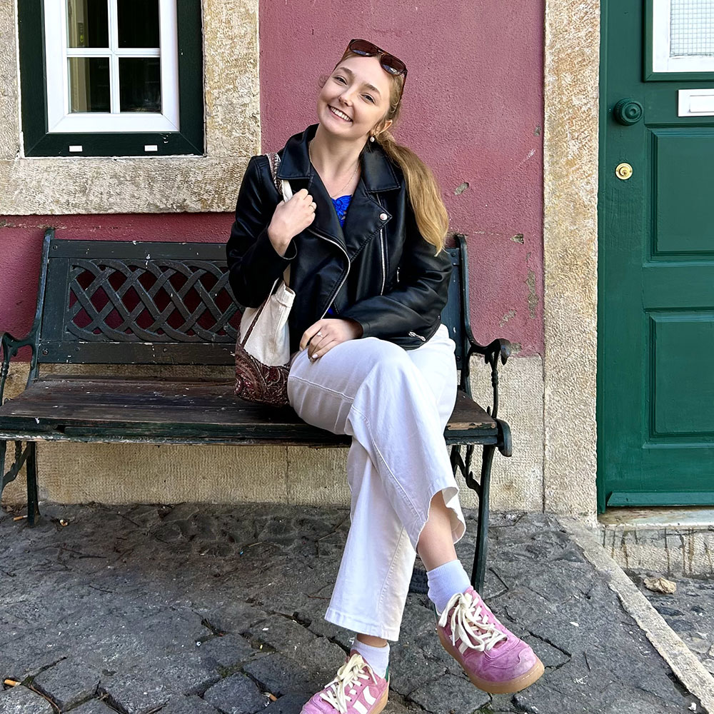 A smiling Alie McDougall sits on a green bench. She is wearing a dark blue shirt, white pants. and pink walking shoes.
